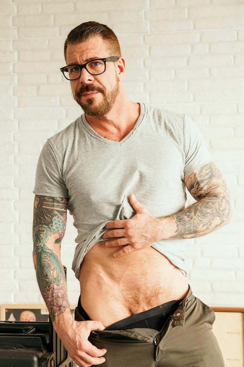 A mature and muscular male model with salt-and-pepper hair and extensive tattoos, reflecting the rugged and professional presence of Rocco Steele.